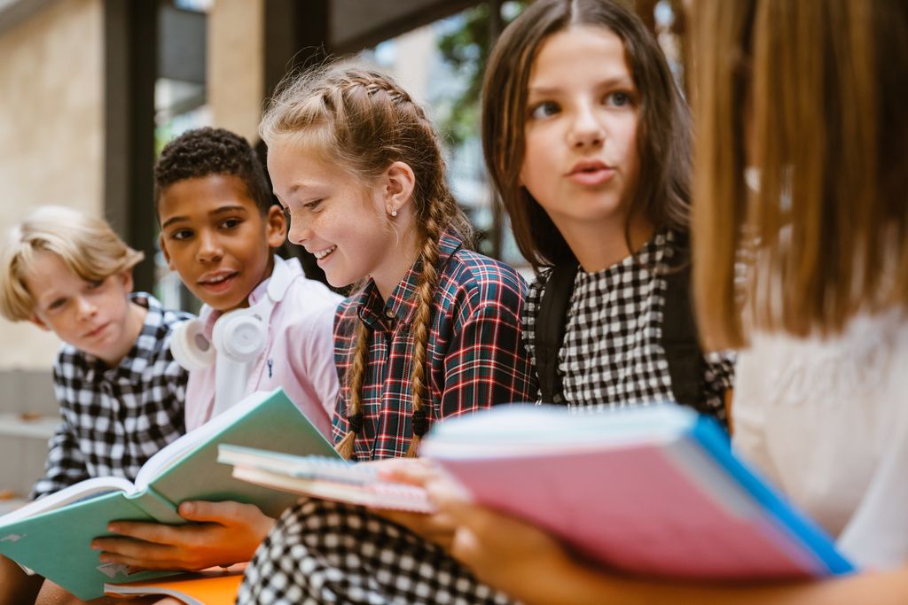 Multiracial pupils doing homework while sitting together by scho Zdjęcie piątki dzieci w wieku wczesnonastoletnim, rozmawiających ze sobą.